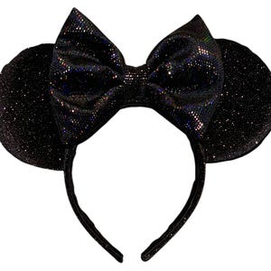 May include: A black headband featuring two circular ears and a large central bow. The entire surface of the headband, ears, and bow is covered in a fine, dark, shimmering glitter material that reflects light with subtle iridescent highlights.