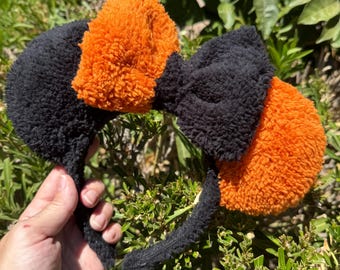 May include: Black headband with orange and black fluffy ears and a bow. The ears are a bright orange color, and the bow is black. The headband is held in a hand against a background of green foliage.