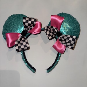 May include: A pair of teal sequined mouse ear headbands. Each ear has a pink satin bow and a black and white checkered bow. The headband is black with teal sequins.