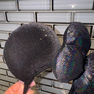 May include: A pair of black mouse ears featuring a glittery fabric finish on the circular ears and a central bow covered in iridescent, multi-coloured reflective sequins that catch the light to create a shimmering, rainbow-like effect.
