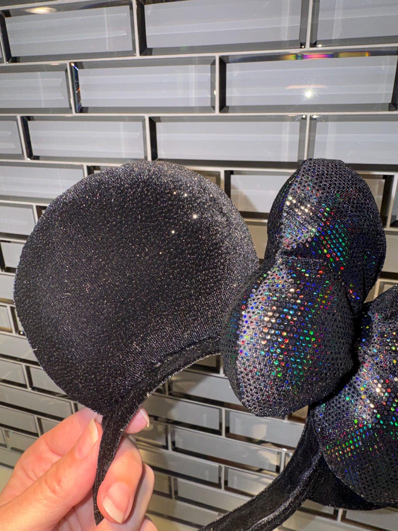 May include: A pair of black mouse ears featuring a glittery fabric finish on the circular ears and a central bow covered in iridescent, multi-coloured reflective sequins that catch the light to create a shimmering, rainbow-like effect.