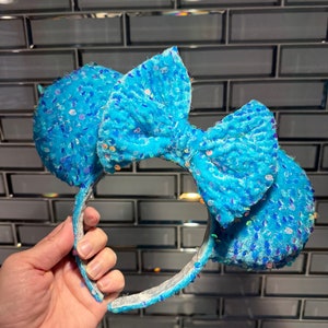 May include: Light blue sequined Minnie Mouse ears with a large bow. The ears are made of a soft, plush material and are decorated with iridescent sequins.
