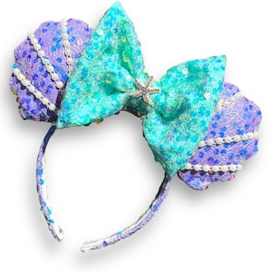 May include: Purple and teal sequined Minnie Mouse ears with a pearl trim and a silver starfish bow.