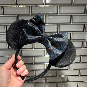 May include: A headband featuring two round black ears covered in a fine glitter texture. A large, central bow sits between the ears, crafted from a black fabric with a holographic, multi-coloured reflective dot pattern that catches the light.