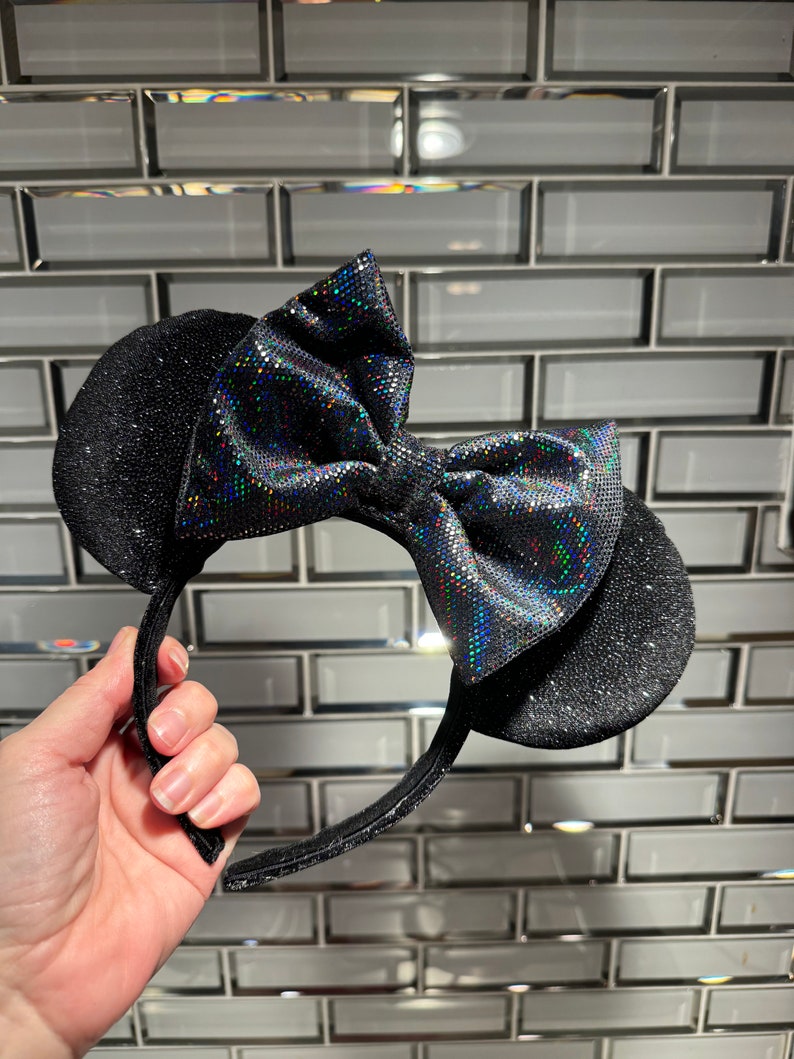 May include: A headband featuring two round black ears covered in a fine glitter texture. A large, central bow sits between the ears, crafted from a black fabric with a holographic, multi-coloured reflective dot pattern that catches the light.
