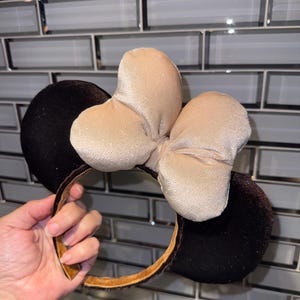 May include: A headband featuring two large, round, black velvet ears and a central, oversized, shimmering champagne-colored fabric bow. The inner band is lined with a metallic gold material, providing a contrast to the dark velvet exterior.