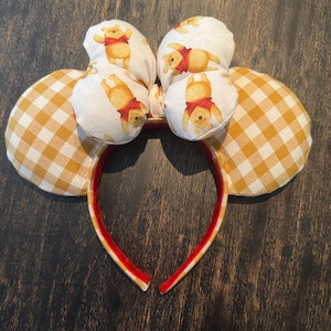 May include: A pair of mouse ears featuring a red headband with yellow trim. The ears are made of yellow and white gingham fabric and white fabric printed with Winnie the Pooh. A fun accessory for fans of the classic character.