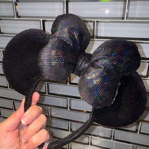 May include: A pair of black headband ears featuring a large, central bow. The ears are covered in a fine, dark glittery texture, while the bow is made of a reflective, iridescent material that displays a multi-coloured, holographic shimmer.