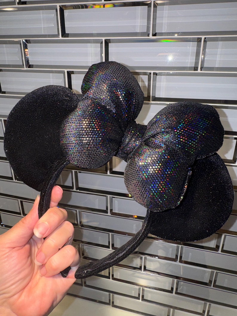 May include: A pair of black headband ears featuring a large, central bow. The ears are covered in a fine, dark glittery texture, while the bow is made of a reflective, iridescent material that displays a multi-coloured, holographic shimmer.