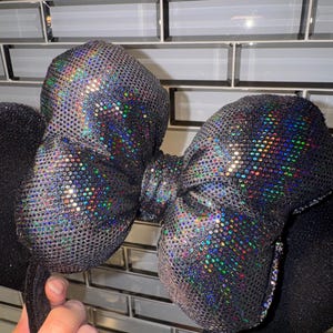 May include: A headband featuring two round black ears and a large central bow. The bow is covered in a dark, reflective material with a holographic, multi-coloured glitter effect that catches the light, creating a shimmering, textured appearance.
