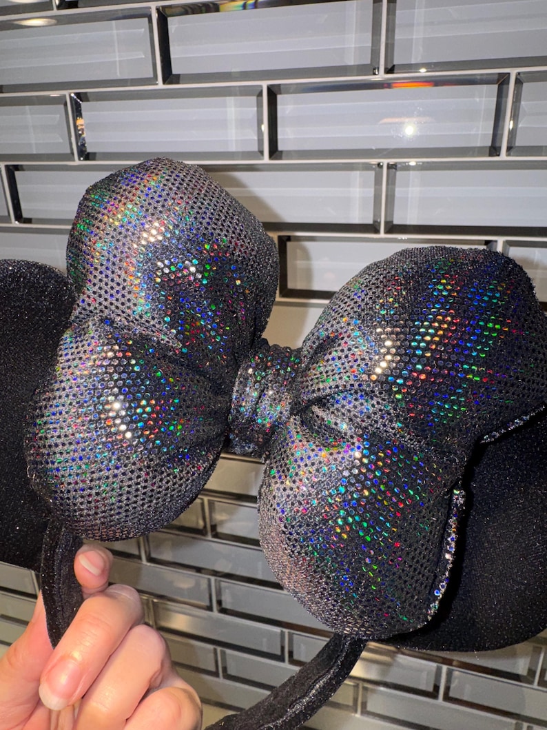 May include: A headband featuring two round black ears and a large central bow. The bow is covered in a dark, reflective material with a holographic, multi-coloured glitter effect that catches the light, creating a shimmering, textured appearance.