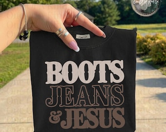 May include: Black sweatshirt with the words "BOOTS JEANS & JESUS" in a layered, neutral color design. The sweatshirt is held up by a hand with rings and a bracelet. The background is blurred with greenery.