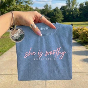 Può includere: Una maglietta blu con le parole "she is worthy" in rosa e "PROVERBI 31" sotto. La maglietta è tenuta in mano da una mano con anelli. Un ornamento circolare trasparente con testo è appeso a una catena.