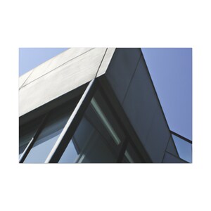 May include: An architectural photograph of a modern building's corner against a clear blue sky. The building features a gray, geometric design with large windows reflecting the sky. The composition emphasizes sharp angles and clean lines.