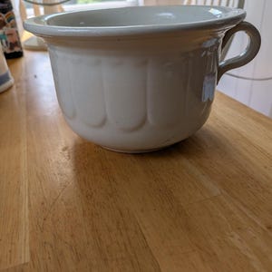 May include: A cream-colored, ceramic chamber pot with a handle. The pot has a wide rim and a rounded body with vertical, scalloped detailing. It sits on a wooden surface.