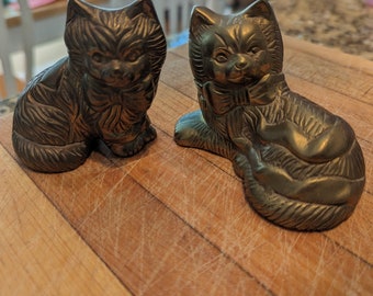 May include: Two brass cat figurines with detailed fur textures and bow ties. One cat is sitting upright, while the other is in a reclining position. Both figures feature a metallic finish with dark patina accents, showcasing intricate facial features.