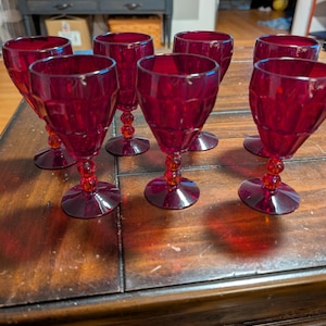 May include: A set of eight ruby red wine glasses. Each glass features a faceted bowl, a beaded stem, and a circular base. The glasses are arranged on a dark wooden table, showcasing their rich color and design.
