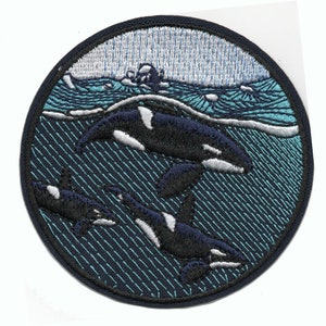 May include: Embroidered patch featuring three orcas swimming in a blue and white ocean. The orcas are black and white with white bellies. The patch is circular and has a blue border.