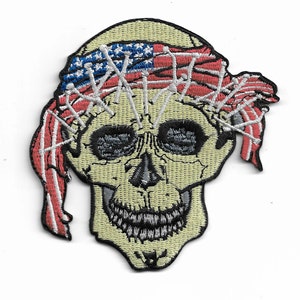 May include: An embroidered patch featuring a white skull with a red, white, and blue bandana over its eyes. The bandana is patterned with stars and stripes, resembling the American flag.