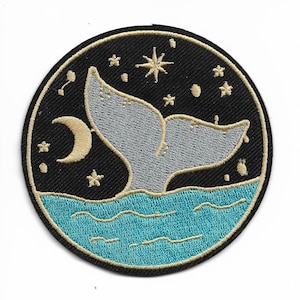 May include: A round embroidered patch with a gray whale tail splashing in turquoise water. The background is black with a gold crescent moon and stars.