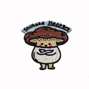 May include: Iron-on patch featuring an embroidered cartoon mushroom with a face and arms crossed. The mushroom is wearing a white cap with the text "Shiitake Happens" in blue.