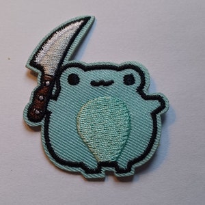 May include: A green frog holding a silver knife with a brown handle. The frog is embroidered on a fabric patch.