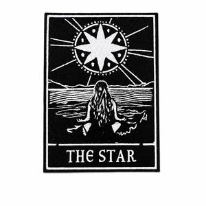 Peut inclure: Un écusson brodé rectangulaire présentant un motif noir et blanc d'une personne assise en position de méditation au bord de l'eau sous une grande étoile à huit branches. Le texte THE STAR est imprimé en blanc au bas de l'écusson.