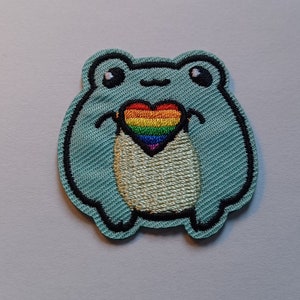 May include: A light blue embroidered frog holding a rainbow heart. The frog has black eyes and a small smile.
