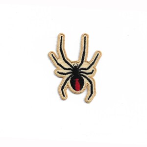 Peut inclure: Patch brodé en forme d'araignée noire et rouge avec un fil beige qui délimite le corps et les pattes de l'araignée.