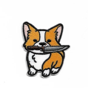 May include: Embroidered patch of a cartoon corgi dog with a knife in its mouth. The corgi is orange and white with black eyes and a black nose. The knife is silver with a black handle.
