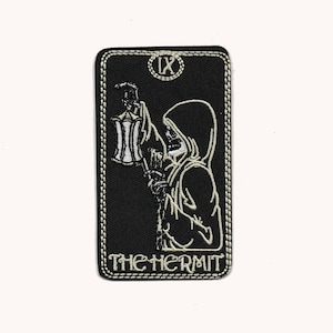 May include: Black and white embroidered patch of the tarot card The Hermit. The card features a hooded figure holding a lantern and the Roman numeral IX.
