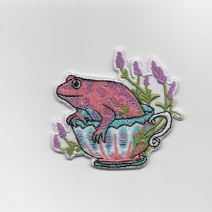 May include: Pink frog sitting in a teacup with lavender flowers around it. The teacup is decorated with pink, blue, and green floral patterns.