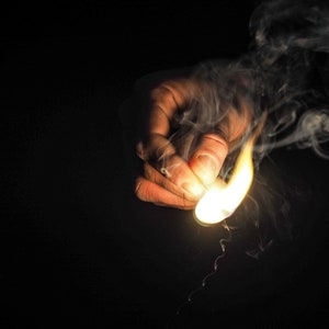 May include: A hand holding a lit match with smoke rising from it against a black background.