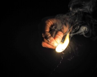 May include: A hand holding a lit match with smoke rising from it against a black background.