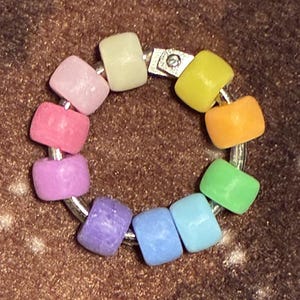 May include: A colorful bracelet featuring a ring of small, square beads in a rainbow of colors. The beads are arranged in a circular pattern, with a silver-colored metal clasp. The bracelet is set against a brown background.