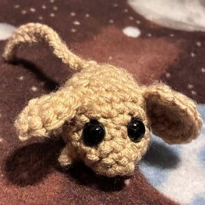 Op de afbeelding: Een handgemaakte, beige gehaakte muis met grote zwarte ogen en flaporen. De muis heeft een lange staart en is gemaakt met een getextureerd garen. Het speelgoed ligt op een bruin en blauw oppervlak.