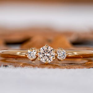 Może przedstawiać: Złoty pierścionek z trzema okrągłymi diamentami. Środkowy diament jest nieco większy od dwóch bocznych kamieni. Pierścionek ma cienką, wypolerowaną obrączkę. Tło jest rozmyte.
