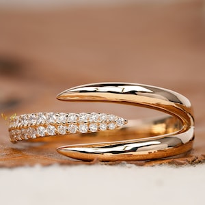 May include: A gold ring with a unique design featuring two curved bands that intersect. One band is set with a row of small diamonds.