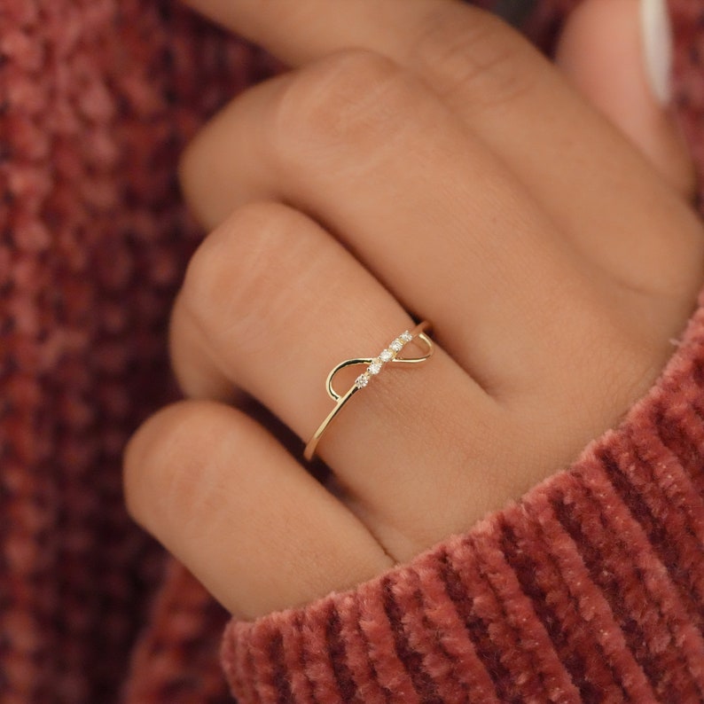K&ouml;nnte beinhalten: Ein goldener Ring mit einem zarten Unendlichkeits-Symbol-Design, das mit kleinen, klaren Steinen verziert ist.