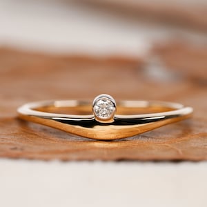 Puede incluir: Un delicado anillo de banda de oro con un solo diamante pequeño y redondo engastado en el centro. La banda está ligeramente curvada hacia arriba, creando un diseño único y moderno.