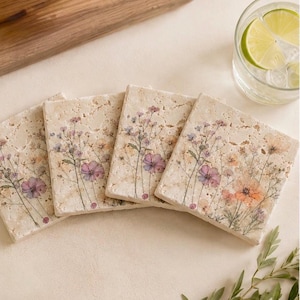 May include: Four square travertine stone coasters featuring a printed design of delicate wildflowers in shades of pink, purple, and orange. The natural porous texture of the beige stone is visible across the surface of each individual coaster.