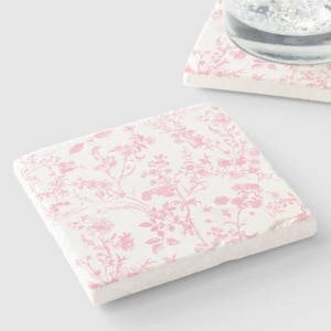 May include: Two square pink floral coasters made of stone. The coasters have a white background with a pink floral pattern. One coaster has a glass with ice and a lime wedge on top.