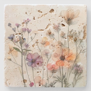 May include: A beige stone coaster with pressed flowers in shades of pink, orange, and white. The flowers are arranged in a random pattern and appear to be pressed between two layers of stone.