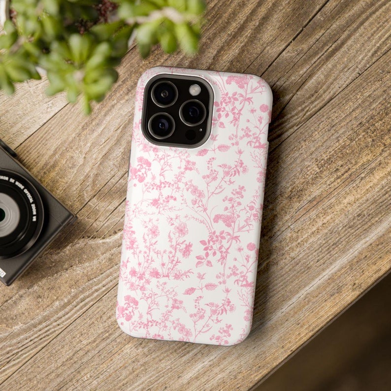 Pode incluir: Uma capa de iPhone branca com um padr&atilde;o floral rosa. A capa tem uma protuber&acirc;ncia de c&acirc;mera elevada e um recorte para a porta de carregamento.