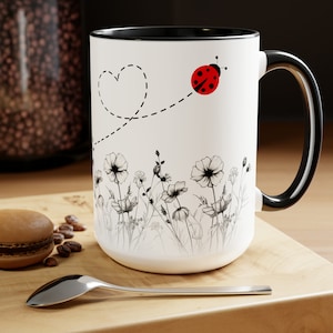 May include: White ceramic mug with a black rim and handle. The mug features a black and white illustration of wildflowers and a red ladybug flying in a dotted line heart shape.