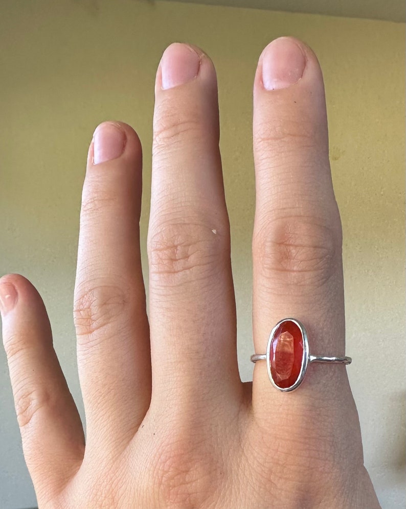 Peut inclure: Une bague en argent avec une pierre ovale rouge. La bague est sur le doigt d'une personne.