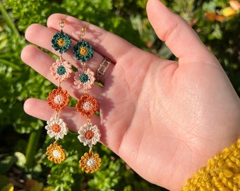 Pode incluir: Um par de brincos longos e pendentes com pingentes de flores de crochê em tons de verde, laranja, branco e amarelo. Os brincos são enfiados em uma corrente dourada.