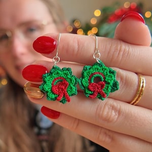 Op de afbeelding: Gehaakte kerstkrans oorbellen. Deze feestelijke oorbellen hebben een groen kransontwerp met een rode strik. De oorbellen worden getoond tegen een achtergrond van een kerstboom en vakantiedecoraties.