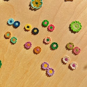Könnte beinhalten: Eine Ansammlung bunter, gehäkelter Blumenapplikationen in verschiedenen Größen und Farbtönen, darunter Blau, Gelb, Rosa, Orange und Grün. Die Blumen sind auf einer hellbraunen Holzoberfläche verstreut und erzeugen eine fröhliche, handgemachte Ästhetik.