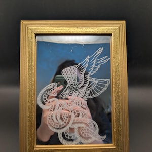 May include: A rectangular mirror in a gold-coloured frame. The mirror has an etched design of an eagle in flight, fighting a serpent. The artwork is detailed, with the figures rendered in white against a blue background. The mirror is supported by a small stand.
